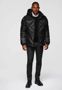 Giacca a pallone nera con cappuccio, caratterizzata da un design trapuntato, chiusura frontale a scatto e polsini elasticizzati. Indossata sopra un maglione nero e jeans.