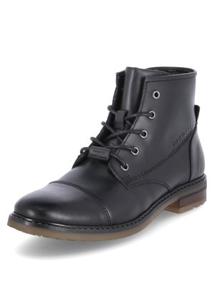 Bota de cuero negro a la altura del tobillo con punta redondeada, cierre con cordones, ojales de metal y suela marrón en contraste. Presenta detalles de costura visibles.