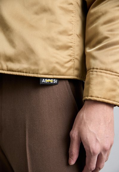 Tan quilted jacket with a smooth texture, featuring a black label reading "ASPESI." Brown pants partially visible, with a soft, matte finish.