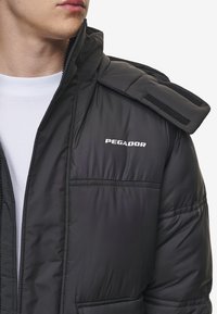 Melna pūkaina jaka ar augstu apkakli, rāvējslēdzēja aizdari un Velcro plecu siksnu. "PEGADOR" logo baltā krāsā uz krūšu.