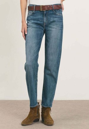 Persoon die blauwe denim jeans draagt met een bruine leren riem en bruine suède enkellaarsjes, staand op een effen vloer.