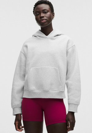 Eine junge Person trägt einen hellgrauen Hoodie mit Fronttasche und leuchtend magentafarbenen Radsportshorts vor einem einfarbigen Hintergrund.