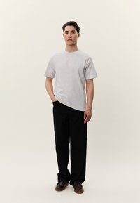 Mladý muž stojaci, oblečený v jednofarebnom svetlosivom tričku, čiernych nohaviciach a hnedých topánkach, s jednou rukou v vrecku na jednoduchom pozadí.