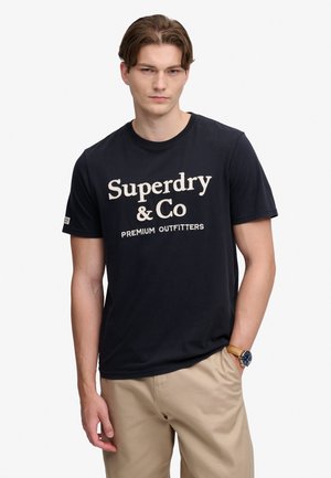 Junger Mann trägt ein schwarzes "Superdry & Co Premium Outfitters" T-Shirt, beige Hose und eine Armbanduhr, steht mit einer Hand in der Tasche.