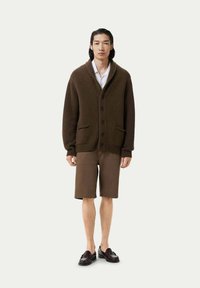 Brun stickad cardigan med sjalkrage, knappstängning och framfickor, i kombination med bruna shorts och mörka skor.