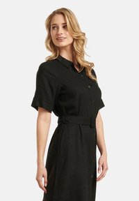 Schwarzes Leinenkleid mit kurzen Ärmeln, durchgehender Knopfleiste und tailliertem Gürtel. Es hat einen Kragen und eine glatte Textur.
