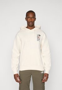 Kremowy hoodie z kieszenią z przodu, z graficznym nadrukiem na lewej piersi. Rękawy i dół są ściągnięte dla lepszej struktury.