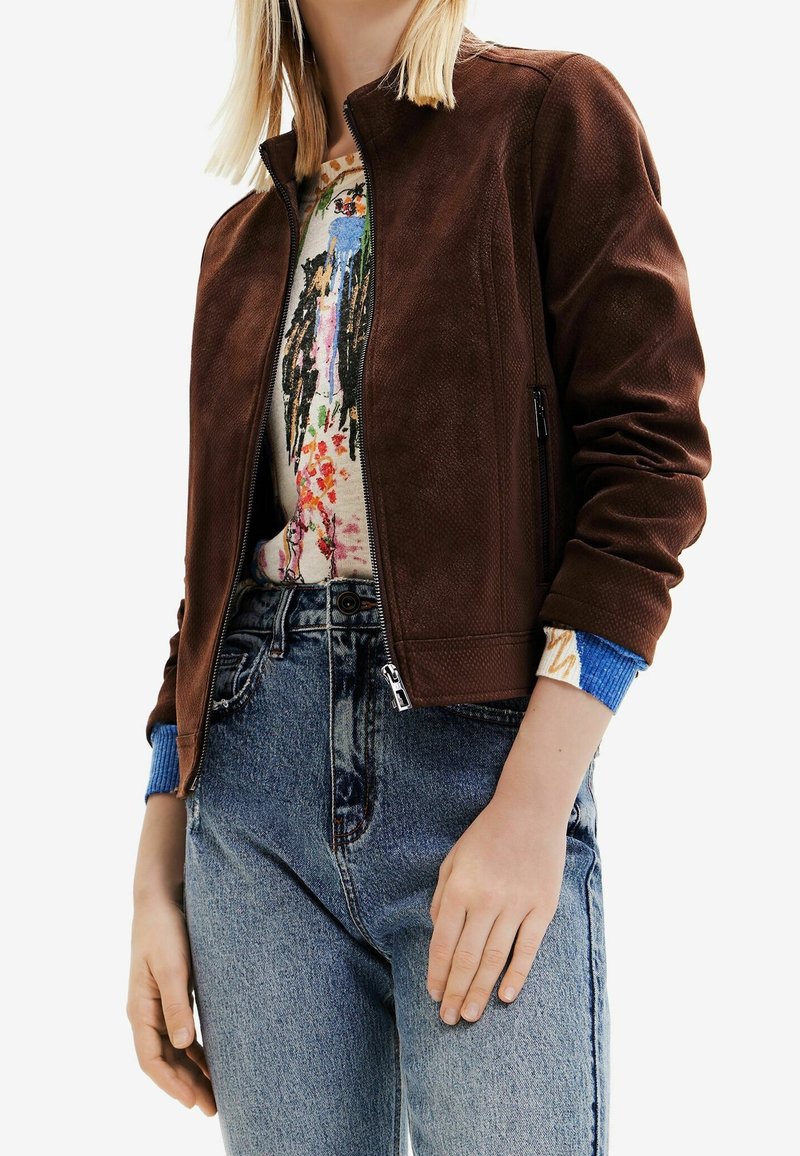 Veste bomber en daim marron avec fermeture éclair avant, poignets côtelés bleus à motifs, portée sur un tee-shirt graphique coloré et un jean en denim.