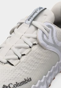 Fehér Columbia sportcipő közeli képe, amely fonott fűzőket, texturált kötött anyagot, a nyelv húzófülén található logót és hullámos talpbetét dizájnt mutat.