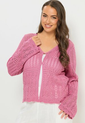 Mujer con cabello largo y castaño, vistiendo un cárdigan de punto rosa sobre una camiseta blanca, sonriendo y posando frente a un fondo liso.