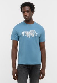 T-shirt bleu à manches courtes avec un graphique de cheval blanc et le texte "MUSTANG", fabriqué en coton avec un col rond et une coupe standard.