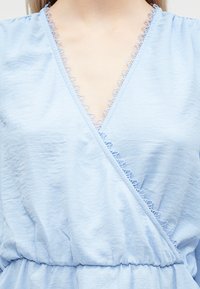 Lyseblå slå-om-top i blødt stof med V-udskæring med fin blondekant og taljeforstærkning for facon.
