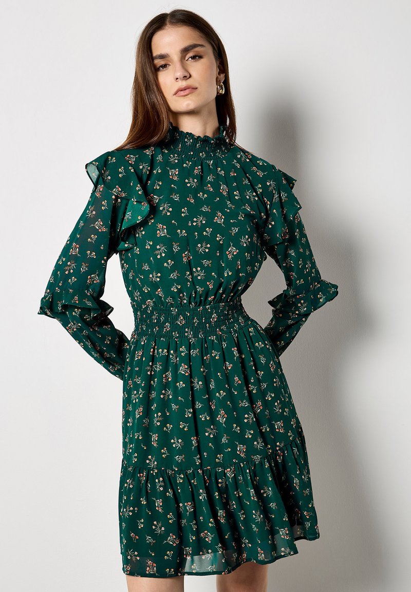 Zielona sukienka w kwiaty z długimi falbaniastymi rękawami i marszczoną talią. Oferuje wielowarstwową spódnicę z małymi nadrukami kwiatowymi na całej powierzchni.