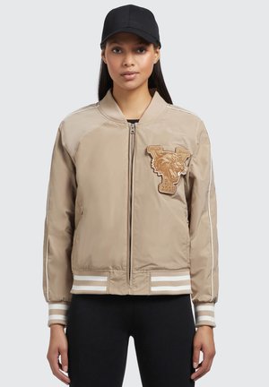 Veste bomber beige avec fermeture éclair, poignets rayés et un logo de panthère brodé brun. Texture lisse avec un tissu uni.