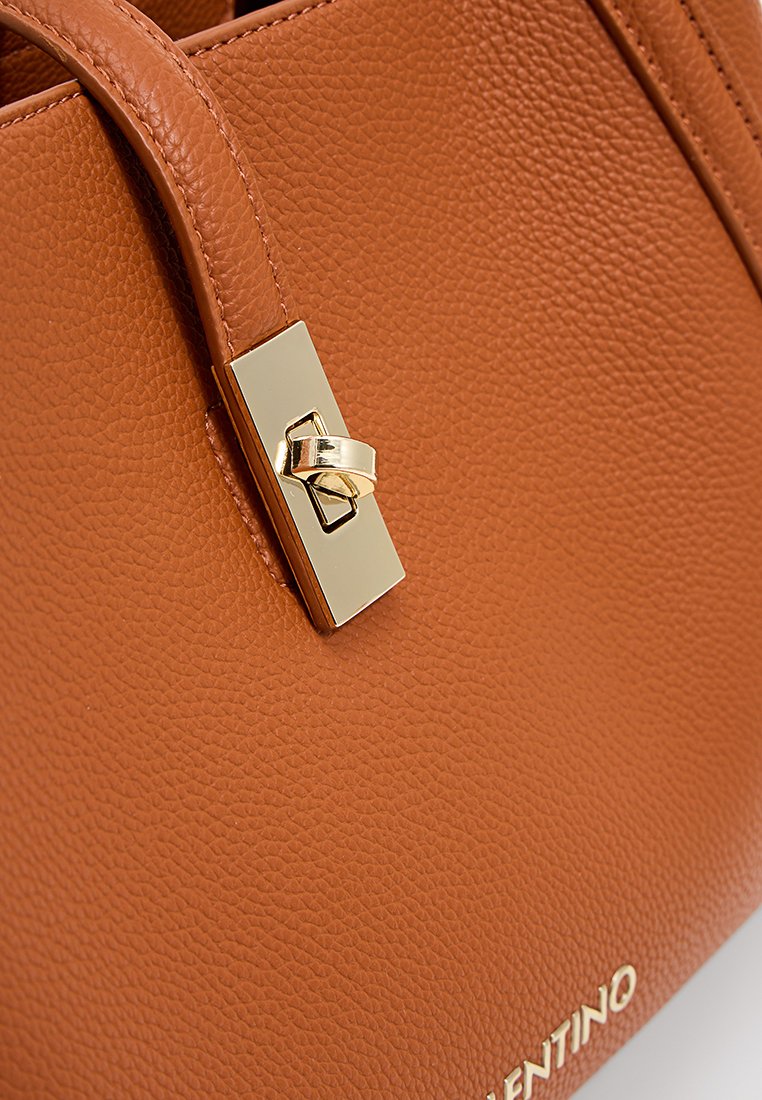 Sac à main en cuir tan avec une surface texturée, doté d'un fermoir et d'accents dorés. Le nom de la marque est embossé en doré au fond.