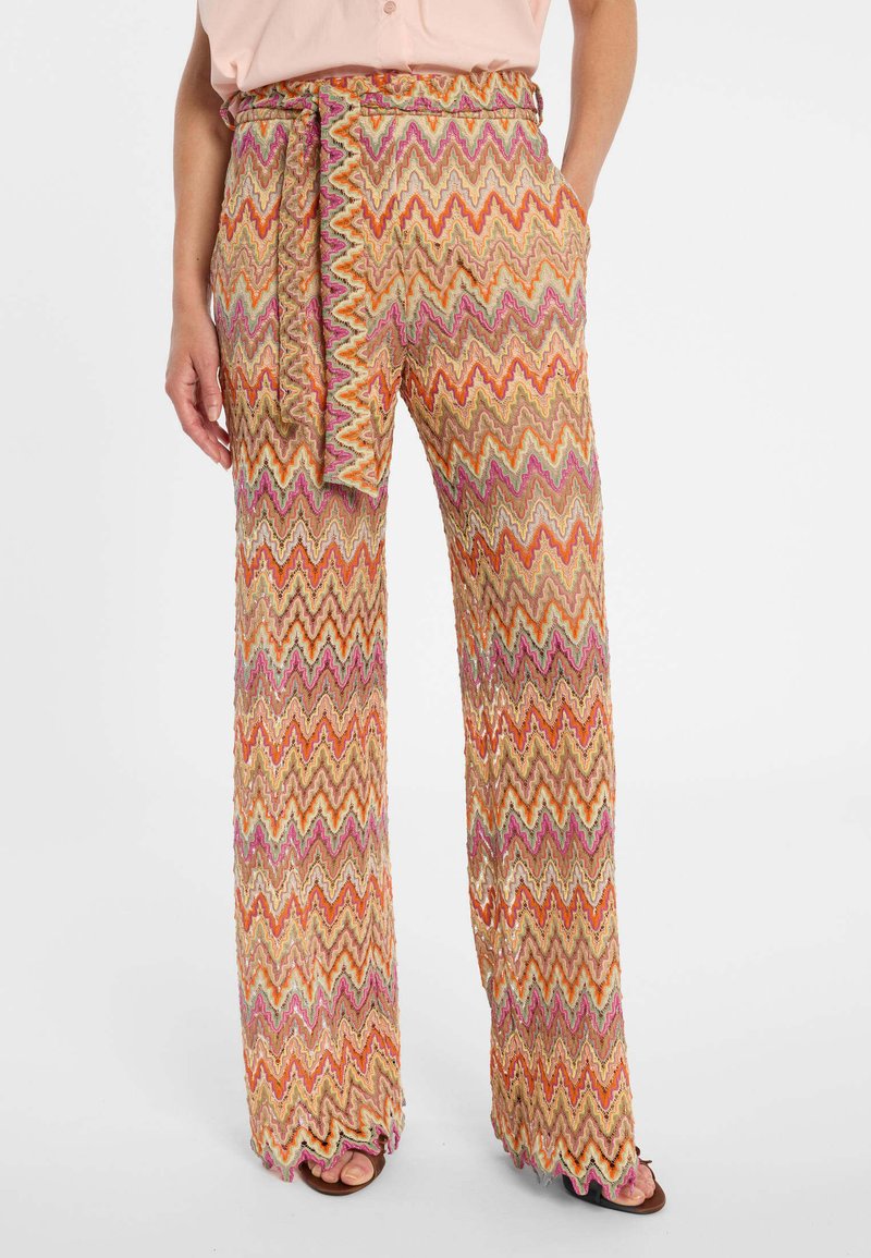 Vrouw die hoge taille wijde pijpenbroek draagt met multicolor zigzagpatroon en ceintuur, gecombineerd met lichtroze top en open sandalen.