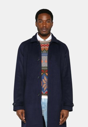 Manteau en laine marine avec un col, des boutons et une texture lisse, porté par-dessus un pull à motifs colorés et un jean bleu clair.