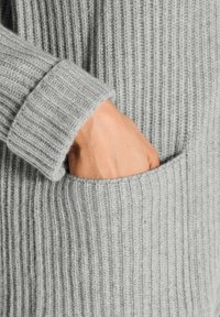 Eine Hand, die in die Tasche eines grauen gerippten Strickpullis gesteckt ist, zeigt die detaillierte Textur und die Manschettennaht.