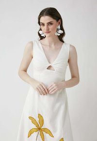 Weißes Kleid mit V-Ausschnitt und einem frontalen Ausschnitt. Verziert mit gelber Palmblattstickerei und aus einem strukturierten Stoff gefertigt. Große weiße Blumenohrringe.