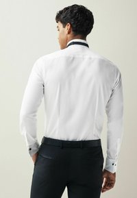 Weißes Langarmhemd mit schwarzen Kragenakzenten. Figurumspielender Schnitt, glattes Material. Getragen mit schwarzen Hosen und einem strukturierten Gürtel.