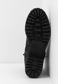 Semelle de botte noire avec un motif de bande de roulement profond et robuste, reposant verticalement contre un angle blanc entre un mur et le sol.