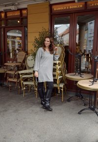 Šedý oversized svetr s výstřihem do V, černé přiléhavé kalhoty a černé kozačky nad kolena. V pozadí se stohují kavárenské židle a stoly.