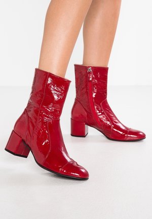 Bottes rouges brillantes mi-mollet avec talons blocs et fermetures éclair latérales portées sur jambes nues, debout sur une surface blanche lisse.