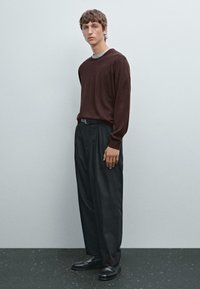 Maglietta a maniche lunghe color mogano con collo grigio abbinata a pantaloni neri larghi. Il modello è in piedi su una superficie scura contro uno sfondo neutro.
