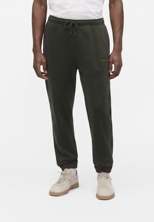 Homme portant un pantalon de jogging vert foncé avec des poignets élastiques et des baskets beiges sur un fond blanc uni.