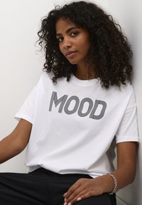 Bijela majica s crnim prugastim natpisom "MOOD." Kratki rukavi, opušteni kroj, nosi se s crnim hlačama, prikazana na jednobojnoj pozadini.
