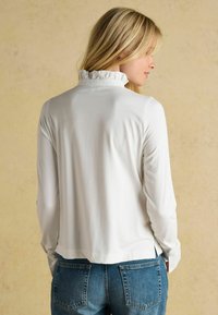 Weißes Langarmoberteil mit einem rüschengeschmückten Kragen, das eine glatte Textur und eine lockere Passform bietet, kombiniert mit blauen Jeans.