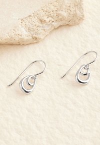 Silberne Ohrhänger mit drei ineinander verschlungenen Tropfenformen. Verfügt über ein poliertes Finish und gebogene Haken. Der Hintergrund ist cremefarben und strukturiert.