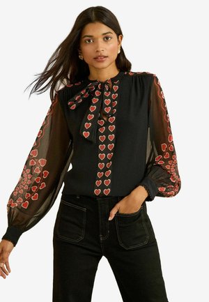 Black blouse with long, sheer sleeves, featuring red heart patterns along the shoulders and center, finished with a neck tie detail.