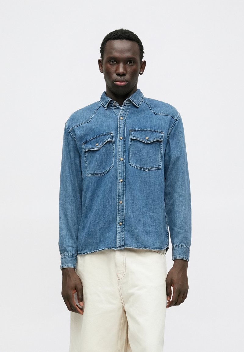 Chemise en denim bleu clair avec des manches longues, un devant boutonné, deux poches sur la poitrine et des coutures sur les épaules et les poignets.