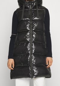 Czarna pikowana kamizelka z wysokim kołnierzem i zamkiem z przodu. Charakteryzuje się błyszczącym wykończeniem, poziomym przeszyciem oraz kieszeniami po bokach.