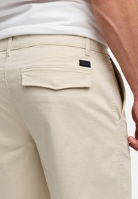 Pantalon en toile beige avec un rabat de poche arrière, des coutures visibles et une petite étiquette noire. Le tissu semble lisse et léger.