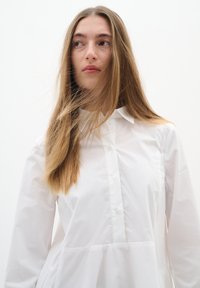 White button-up shirt featuring a collared neckline, long sleeves, and a peplum silhouette. Smooth texture without patterns or accents.