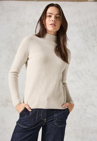 Helle beige gerippte Pullover mit hohem Hals und langen Ärmeln, pr�äsentiert in einer weichen Textur und lockeren Passform. Kombiniert mit dunkler Denim.
