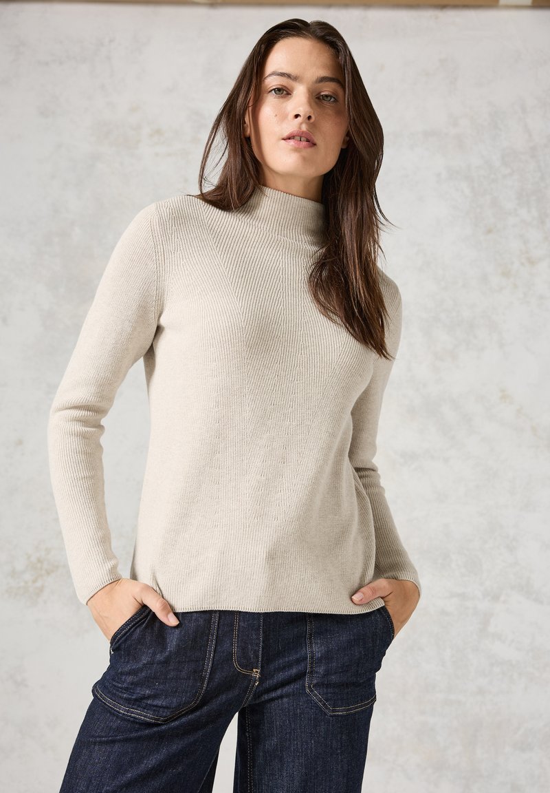 Helle beige gerippte Pullover mit hohem Hals und langen Ärmeln, präsentiert in einer weichen Textur und lockeren Passform. Kombiniert mit dunkler Denim.