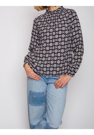 Femme portant un chemisier à manches longues avec un motif géométrique noir et blanc et un jean bleu clair avec des patchs en denim, debout devant un fond uni.