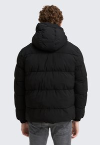 Mand med krøllet hår iført en sort pufferjakke med hætte, stående med ryggen til, iført grå jeans mod en lys baggrund.