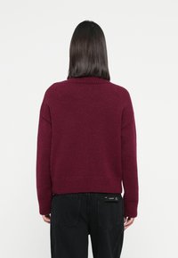 Burgundy, grovstickad tröja med hög krage, ribbstickade ärmslut och nederkant. Bärs över svarta jeans och har en avslappnad passform.