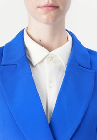 Modrý blazer s klopami, texturovaná tkanina a bílá košile s límečkem pod ním, se zapínáním na knoflíky a hladkým povrchem.