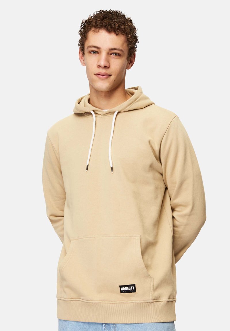 Beige Kapuzenpullover aus weichem Material, mit einer vorderen Tasche, verstellbaren Kordeln und einem kleinen schwarzen Logo-Tag am unteren Ende.