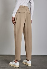 Beige broek met een elastische tailleband, twee achterzakken en tapse pijpen, gecombineerd met witte sneakers op een gestructureerde grijze vloer.