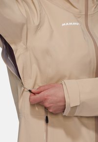 Beige wasserdichte Jacke mit hohem Kragen, verstellbarem Kordelzug an der Taille und mit Reißverschlüssen versehenen Taschen. Sie verfügt über einen glatten, strukturierten Stoff.