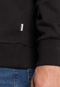 Sudadera negra con puños acanalados, que presenta una pequeña etiqueta de marca blanca. Combinada con unos vaqueros azules, que muestran una textura de tela suave.