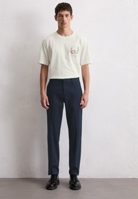 T-shirt blanche à manches courtes avec un design graphique sur la poitrine, associée à un pantalon noir sur mesure et des bottes noires à lacets.