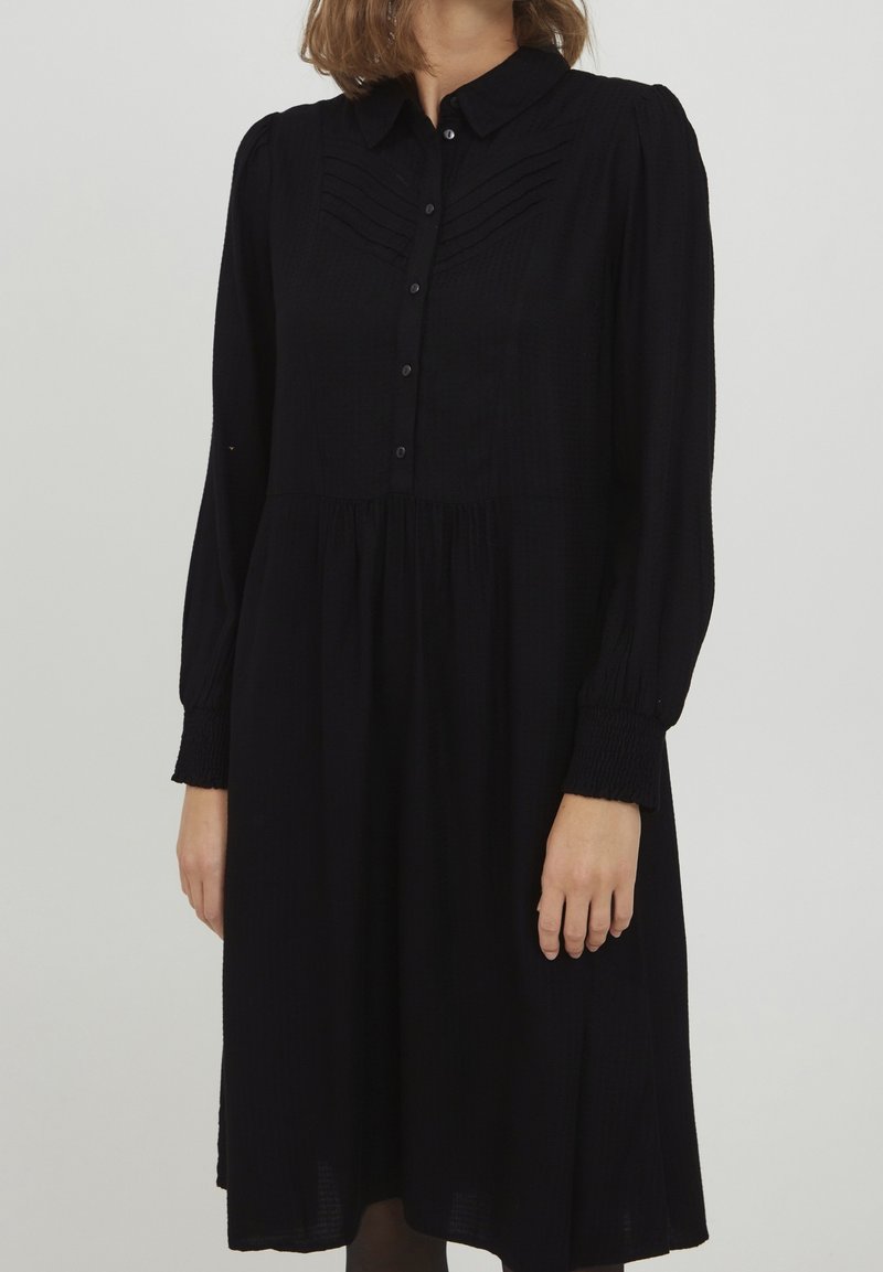 Robe noire avec un col, devant boutonné, manches longues avec des poignets froncés, et un panneau texturé sur le buste. En dessous de la taille, elle tombe doucement.