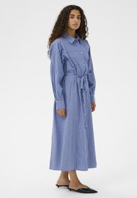 Femme aux longs cheveux bouclés portant une robe-chemise midi à rayures verticales bleues et blanches avec une ceinture à la taille et des chaussures plates noires à bouts pointus.